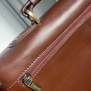 May include: A smooth brown leather bag featuring a top handle attached with antique brass-toned hardware. A horizontal zippered pocket runs across the front, equipped with a matching brown zipper tape and a rectangular metal pull engraved with VISCONTI.