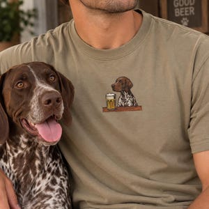 May include: An olive green crew neck t-shirt featuring an embroidered graphic of a brown and white spotted dog sitting at a bar next to a glass of beer. The shirt has a simple design with a small, detailed illustration centered on the chest area.