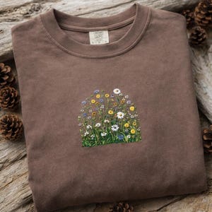 以下が含まれることがあります： 刺繍された花柄が施された茶色のクルーネックのスウェットシャツ。刺繍は、白いデイジー、黄色の花、青いワイルドフラワーなど、さまざまなカラフルな花が、緑の草の基盤に配置されています。