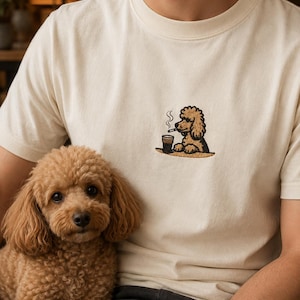 May include: An off-white crew neck t-shirt featuring a small embroidered graphic on the chest. The embroidery depicts a brown poodle sitting at a table with a steaming dark beverage and a cigarette. The shirt has short sleeves and a ribbed collar.
