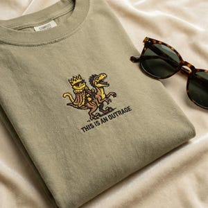 May include: Olive green t-shirt with an embroidered design of two dinosaurs, one wearing a crown. The text "THIS IS AN OUTRAGE" is below the design. Tortoiseshell sunglasses rest on the shirt.