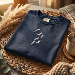 May include: A navy blue t-shirt with an embroidered design of five white birds in flight. The shirt has a crew neck and a tag that reads "Comfort Colors". The shirt is folded on a woven mat with a neutral-toned background.
