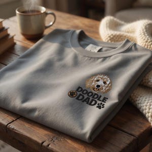 May include: A gray t-shirt with an embroidered doodle dog design and the text "DOODLE DAD". The shirt is folded on a wooden surface, with a cup of coffee and a blanket in the background.