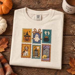 May include: A cream-coloured t-shirt featuring a graphic print of six rectangular postage stamp designs. The illustrations include a jack-o'-lantern, a ghost with a bow, a cauldron, a beagle dog, a haunted house, and a tombstone with a skull.