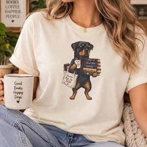 Könnte beinhalten: Ein cremefarbenes T-Shirt mit einer Grafik eines Rottweilers, der eine Sonnenbrille trägt, einen Stapel Bücher und einen Kaffeebecher hält und eine Tragetasche mit der Aufschrift "Libraries Make Life Better" trägt. Ein weißer Becher mit "Good Books Happy Dogs" ist zu sehen.