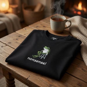 Może przedstawiać: Czarna bluza z ilustracją zielonej i białej żaby siedzącej na stołku oraz napisem "nonsense!" w kolorze białym. Bluza jest złożona na drewnianym stole. W tle paruje filiżanka herbaty.