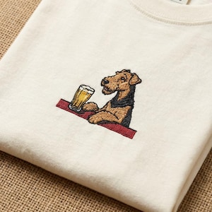 May include: Cream-colored t-shirt featuring an embroidered Airedale Terrier dog with a beer. The dog is brown and black, sitting behind a red bar, with a glass of beer. The shirt is folded.