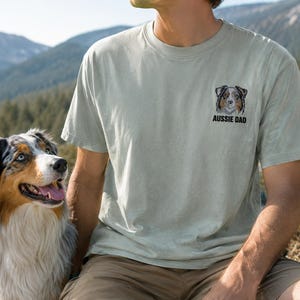 May include: A light sage green short-sleeve t-shirt featuring a small embroidered graphic on the left chest. The graphic depicts a brown, black, and white Australian Shepherd dog face above the black text that reads AUSSIE DAD.