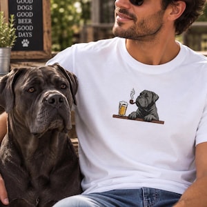 May include: A white t-shirt featuring an embroidered graphic of a dark grey dog resting its paws on a wooden bar counter. The dog is depicted smoking a pipe next to a glass of beer with foam. The design is positioned on the chest area of the shirt.