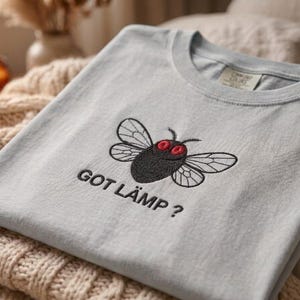 Puede incluir: Una camiseta azul claro con cuello redondo, con un diseño bordado de una mosca con ojos rojos y el texto "GOT LÄMP?". La camiseta está doblada y colocada sobre una manta de punto beige.