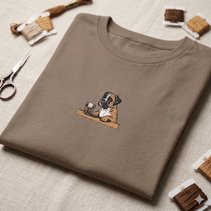May include: A brown t-shirt with an embroidered Boxer dog design. The dog is shown with a pint of beer, resting its paws on a wooden bar. The shirt is folded, with embroidery threads and scissors in the background.