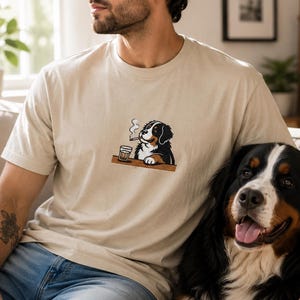 May include: A beige short-sleeved t-shirt featuring a small embroidered graphic on the chest. The graphic depicts a Bernese Mountain Dog sitting at a table with a glass of liquid and a smoking cigarette, rendered in black, white, and brown.