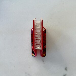 May include: A hair claw clip featuring a vibrant red outer shell with a dark, mottled pattern and a contrasting translucent, pearlescent white inner mechanism with interlocking teeth designed to securely hold hair in place.