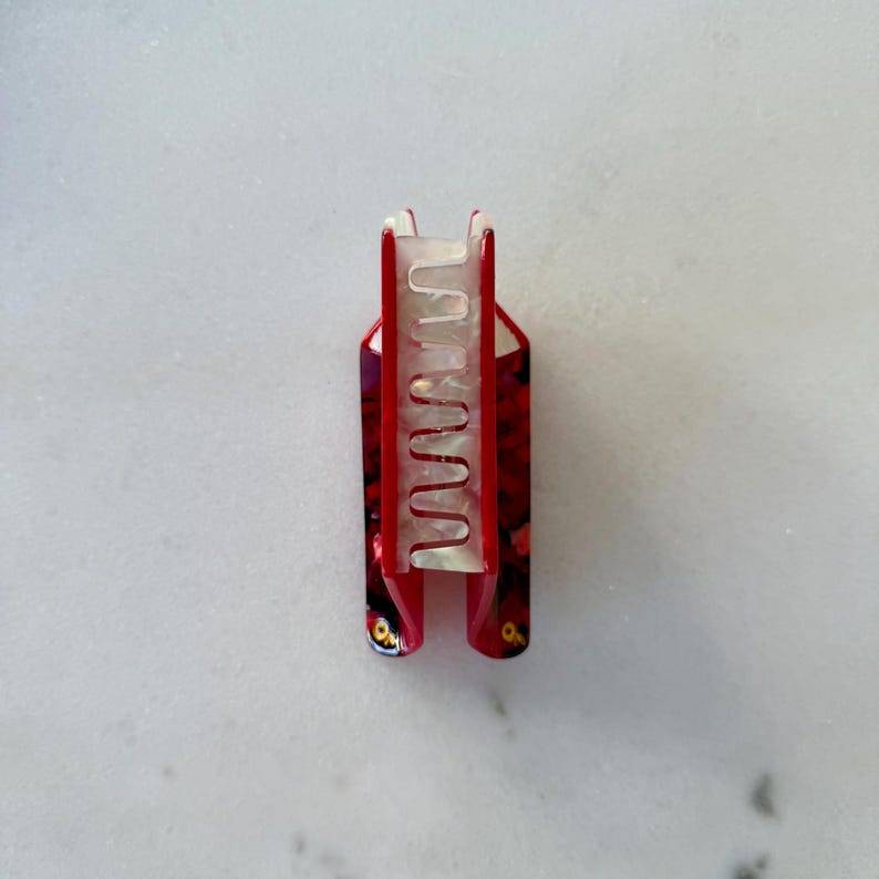 May include: A hair claw clip featuring a vibrant red outer shell with a dark, mottled pattern and a contrasting translucent, pearlescent white inner mechanism with interlocking teeth designed to securely hold hair in place.