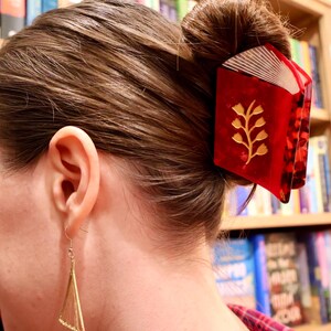 May include: A red hair claw clip shaped like an open book with a gold floral design on the cover and white page details. The clip is secured in a brown hair bun. A gold triangular earring hangs from the earlobe, featuring thin wire strings.