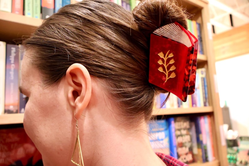 May include: A red hair claw clip shaped like an open book with a gold floral design on the cover and white page details. The clip is secured in a brown hair bun. A gold triangular earring hangs from the earlobe, featuring thin wire strings.