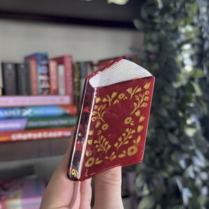 May include: A miniature book ornament with a glossy deep red cover featuring a gold floral vine pattern. The spine is dark red, and the pages are represented by white layered edges. The object is shaped like a small, closed hardcover book.