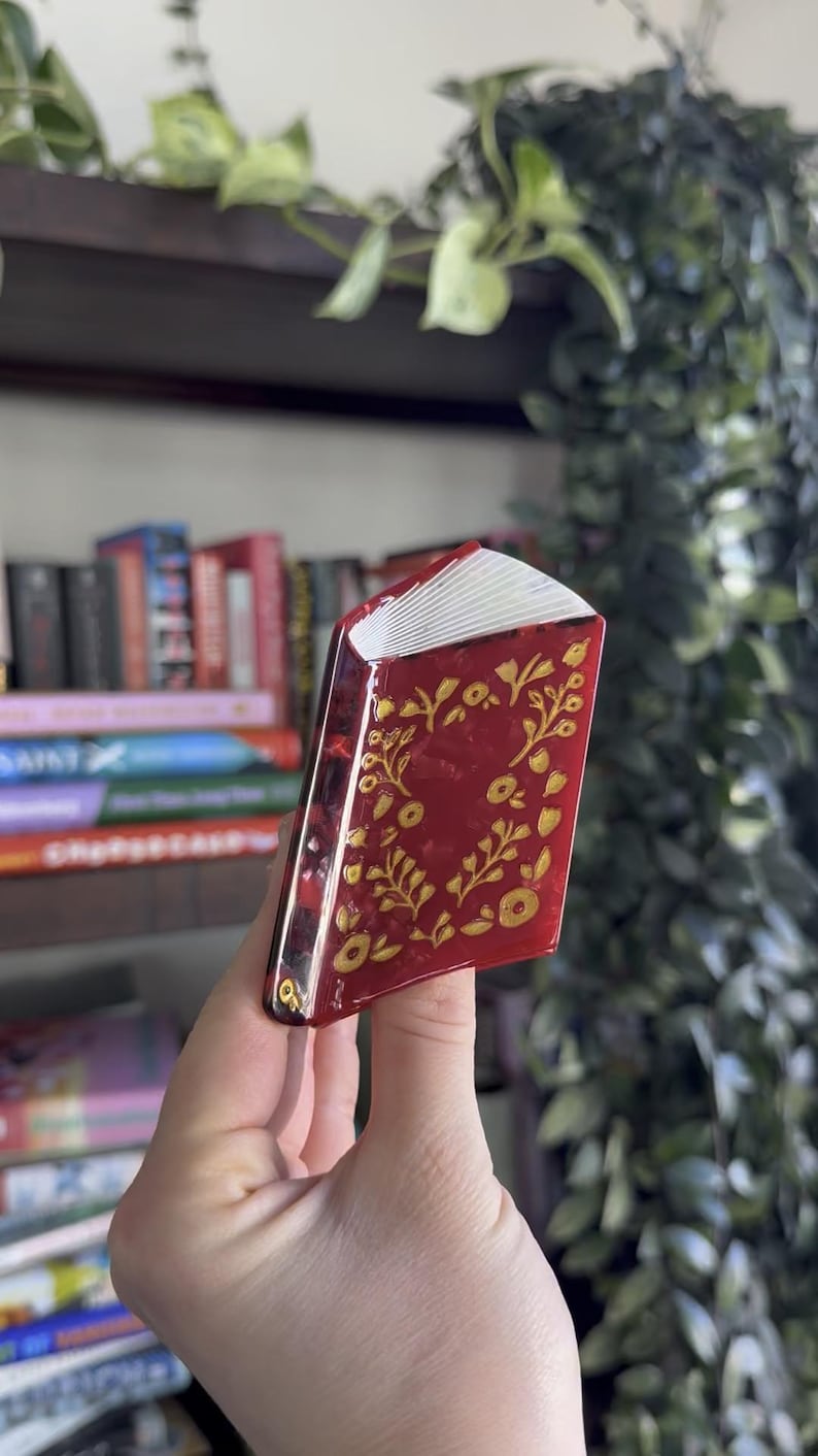 May include: A miniature book ornament with a glossy deep red cover featuring a gold floral vine pattern. The spine is dark red, and the pages are represented by white layered edges. The object is shaped like a small, closed hardcover book.
