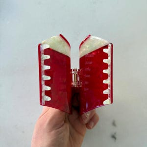 May include: A rectangular hair claw clip featuring a vibrant red marbled pattern with contrasting white pearlescent accents along the top edges and the interlocking teeth. The accessory includes a central metal spring mechanism for opening.