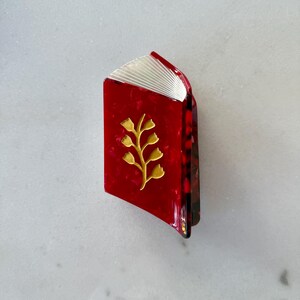 May include: A hair claw clip shaped like an open book with a glossy red marbled finish. The cover features a gold-toned metallic sprig design with small leaves. The top edge shows white, fan-folded pages, and the side reveals a dark tortoiseshell pattern.