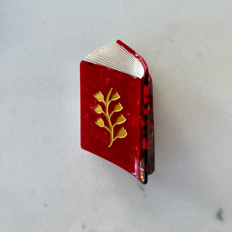 May include: A hair claw clip shaped like an open book with a glossy red marbled finish. The cover features a gold-toned metallic sprig design with small leaves. The top edge shows white, fan-folded pages, and the side reveals a dark tortoiseshell pattern.