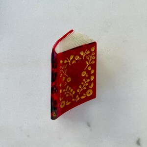 May include: A small, three-dimensional book-shaped pin or charm featuring a glossy red cover with a gold-colored floral pattern. The spine is dark with a mottled texture, and the pages are represented by white, layered, fan-like ridges.