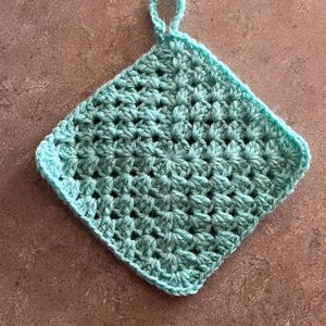 Puede incluir: Un agarrador cuadrado de ganchillo hecho a mano en color verde azulado claro. El agarrador presenta un patrón de puntadas texturizado y un lazo para colgar. El artículo se muestra sobre una superficie marrón.