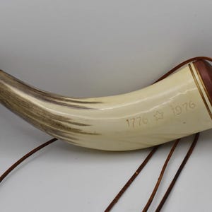 Könnte beinhalten: Ein gebogenes Pulverhorn mit cremefarbenem Körper und brauner Maserung, einem dunkelbraunen Holzsockel und einer goldfarbenen Metallspitze. Der Text 1776, ein Sternsymbol und 1976 sind eingraviert. Eine dünne braune Lederschnur ist befestigt.