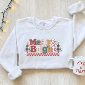 Może przedstawiać: Biała bluza z długim rękawem z napisem Merry & Bright w wielokolorowych blokowych literach, otoczona dwiema czarno-białymi choinkami w kratę, wraz z pasującym białym ceramicznym kubkiem z nadrukiem Merry & Bright.