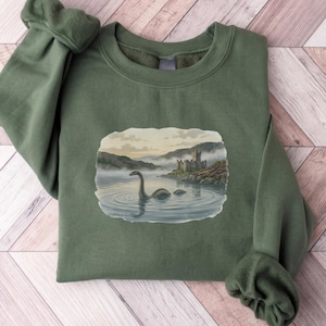 May include: An olive green crewneck sweatshirt featuring a central graphic print of a long-necked creature swimming in a misty lake near a stone castle ruin. The sweatshirt has long sleeves with ribbed cuffs and a ribbed neckline.
