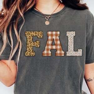 May include: A charcoal grey short-sleeve t-shirt featuring the letters FAL printed across the chest. The letter F has a brown leopard print, the letter A has a brown and white buffalo plaid pattern, and the letter L has a floral print.