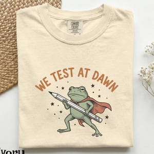 May include: An ivory t-shirt featuring a graphic of a green frog wearing a red cape and holding a large pencil. Above the frog, the text reads WE TEST AT DAWN in brown letters. Small black stars are scattered around the frog on the shirt fabric.