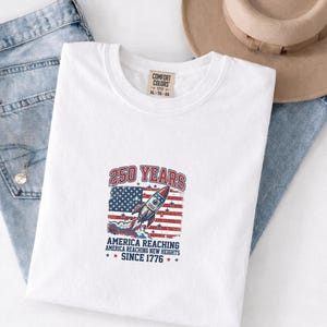 May include: White t-shirt with a graphic commemorating 250 years of America. The design features a rocket, the American flag, and the text "250 YEARS AMERICA REACHING NEW HEIGHTS SINCE 1776". The shirt is size XL-TG-EG.