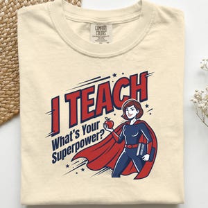 Puede incluir: Una camiseta de color marfil con un gráfico de un personaje de superhéroe con capa, que sostiene una manzana roja y un libro con la etiqueta Knowledge. El texto dice I TEACH What's Your Superpower? en letras rojas y azul marino con estrellas.