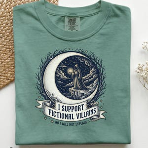 May include: Light green t-shirt with a graphic featuring a cloaked figure on a crescent moon, holding a glowing orb. The design includes the text "I SUPPORT FICTIONAL VILLAINS" and "NO I WILL NOT EXPLAIN". The shirt is made of Comfort Colors material.