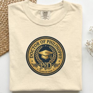 Puede incluir: Camiseta de color marfil con cuello redondo. La camiseta presenta un gráfico circular con las palabras "DOCTOR OF PHILOSOPHY" y "PhD CLASS OF 2026" en dorado y azul marino. Un birrete de graduación está en el centro del gráfico.