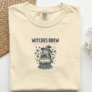 以下が含まれることがあります： 「Witches Brew」の文字と、大釜をかき混ぜる漫画の魔女が描かれたアイボリー色のTシャツ。デザインには星、コーヒー豆、その他の装飾要素が含まれています。このシャツは柔らかい素材で作られています。