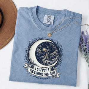 May include: A light blue Comfort Colors t-shirt featuring a graphic of a robed figure holding a glowing orb in front of a crescent moon. The text reads "I SUPPORT FICTIONAL VILLAINS" and "NO I WILL NOT EXPLAIN". A gold necklace and lavender sprig are also visible.