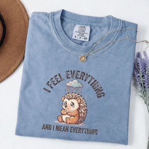 May include: A blue short-sleeve t-shirt featuring a graphic of a crying hedgehog under a cloud with the text I FEEL EVERYTHING AND I MEAN EVERYTHING. The shirt has a crew neckline and a Comfort Colors brand label on the inside collar.