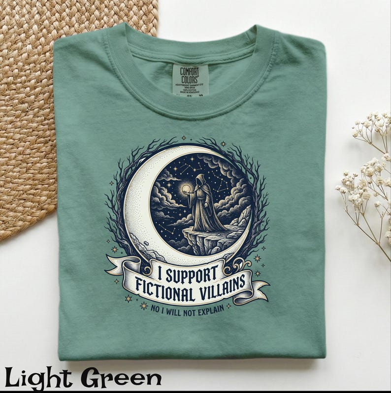 May include: Light green t-shirt with a graphic featuring a cloaked figure on a crescent moon, holding a glowing orb. The design includes the text "I SUPPORT FICTIONAL VILLAINS" and "NO I WILL NOT EXPLAIN". The shirt is made of Comfort Colors material.