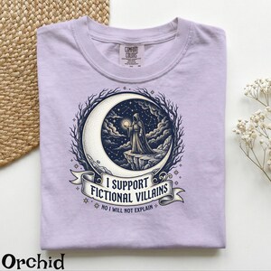 May include: Orchid-colored t-shirt with a graphic of a robed figure holding a glowing orb, standing on a crescent moon. The design includes the text "I SUPPORT FICTIONAL VILLAINS" and "NO I WILL NOT EXPLAIN".