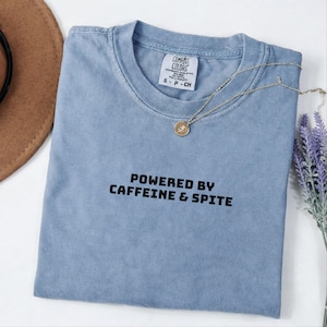 以下が含まれることがあります： クルーネックの半袖ブルーTシャツで、内側の襟元にComfort Colorsのブランドラベルが付いています。胸の中央には、太字のサンセリフ体で「POWERED BY CAFFEINE & SPITE」という黒いテキストがプリントされています。