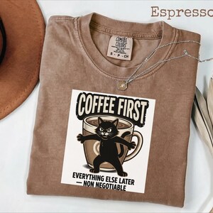 May include: A brown short-sleeved t-shirt featuring a graphic of a black cat standing in front of a coffee mug. The text reads COFFEE FIRST EVERYTHING ELSE LATER - NON NEGOTIABLE. The shirt has a crew neckline and a Comfort Colors brand label.