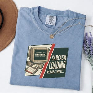 May include: A light blue t-shirt featuring a graphic of a vintage computer monitor with a loading bar at 95 percent and a floppy disk. The text reads SARCASM LOADING PLEASE WAIT. The shirt has a white neck label with the text COMFORT COLORS.