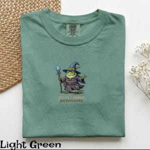 May include: Light green t-shirt featuring a frog wizard graphic. The frog is dressed in a purple robe and a blue hat, holding a staff. The text "OFF TO COMMIT SHENANIGANS" is printed below the frog.
