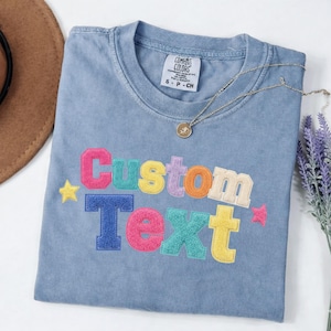 Op de afbeelding: Een blauw t-shirt met de geborduurde tekst Custom Text in kleurrijke letters in varsity-stijl. Het shirt heeft een Comfort Colors merklabel in de nek en is versierd met kleine geborduurde sterren in geel en roze.