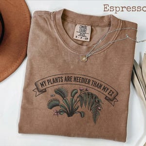 Może przedstawiać: Koszulka w kolorze espresso z grafiką muchołówki i paproci oraz nadrukowanym czarnym napisem "MY PLANTS ARE NEEDIER THAN MY EX". Koszulka posiada okrągły dekolt oraz metkę marki Comfort Colors umieszczoną wewnątrz kołnierza.