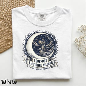 May include: White t-shirt with a graphic featuring a hooded figure holding a glowing orb on a crescent moon, surrounded by stars and the text "I SUPPORT FICTIONAL VILLAINS". The shirt is made of a soft material.