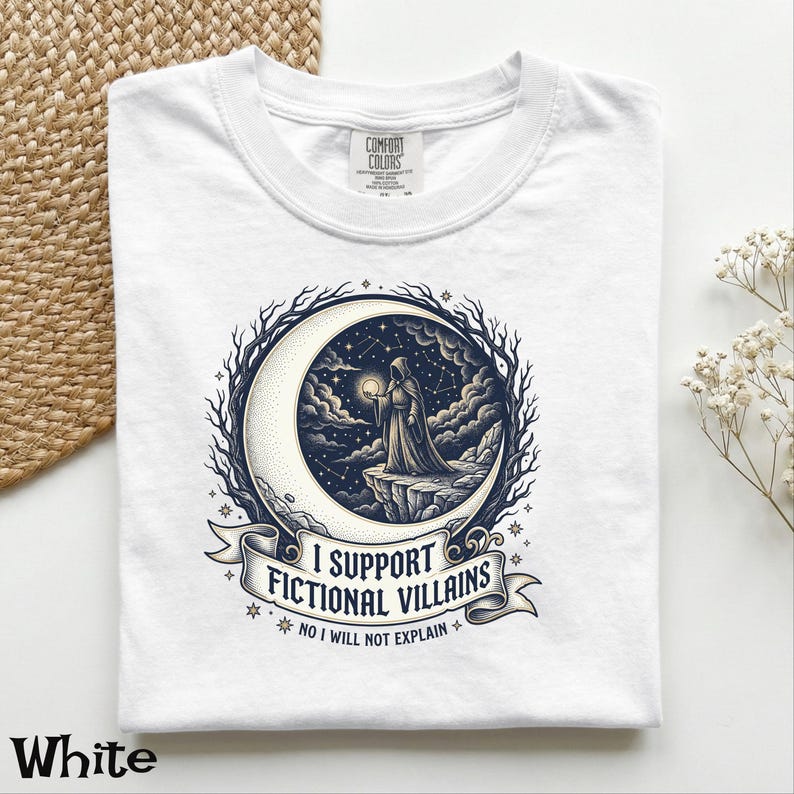 May include: White t-shirt with a graphic featuring a hooded figure holding a glowing orb on a crescent moon, surrounded by stars and the text "I SUPPORT FICTIONAL VILLAINS". The shirt is made of a soft material.