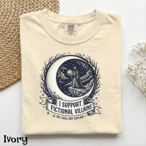 May include: Ivory-colored t-shirt with a graphic featuring a hooded figure standing on a cliff within a crescent moon, surrounded by stars and a banner that reads "I SUPPORT FICTIONAL VILLAINS". The text "NO I WILL NOT EXPLAIN" is below the banner.